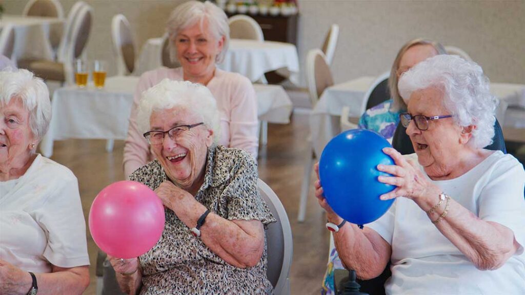 Older adult choosing clothing beside a calendar while a caregiver offers support in a bright apartment